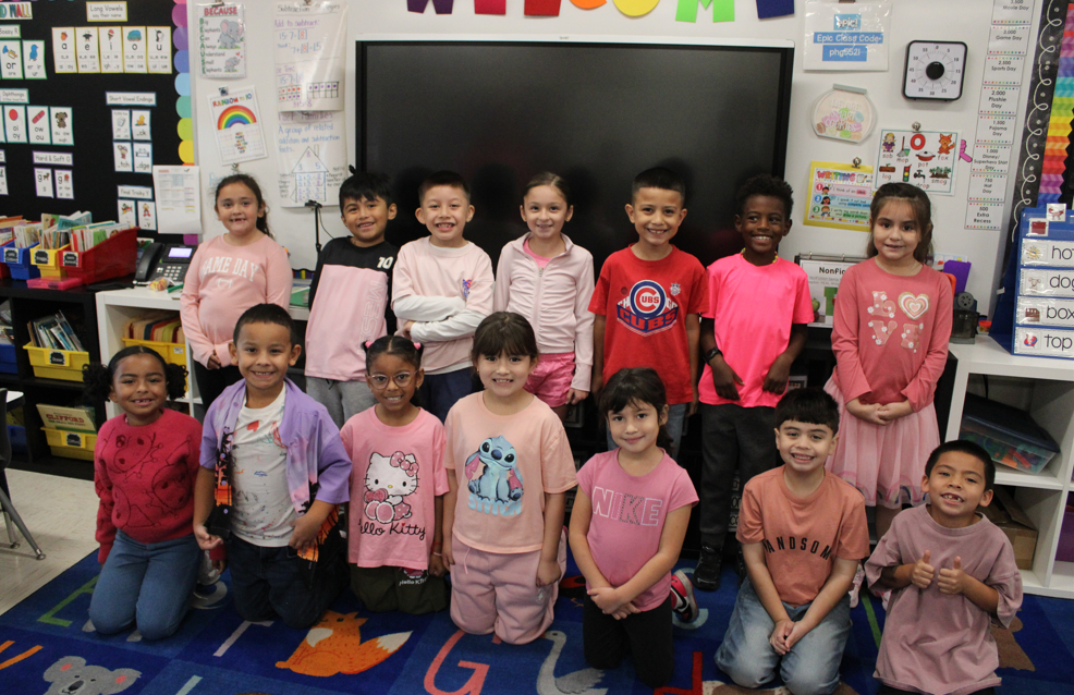A cheerful group of elementary school children pose together in a colorful classroom, with a large blackboard, learning displays, and classroom shelves behind them.