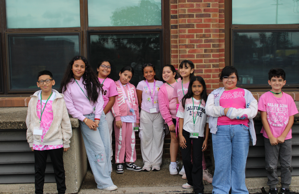 A cheerful group of children in pink casual clothes pose outside a brick school building, smiling beneath large windows on an overcast day.