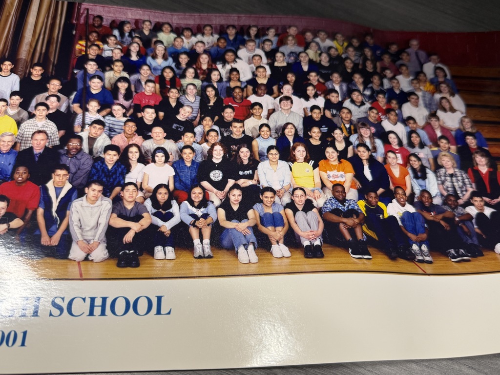 A large, diverse school group poses in rows on an auditorium stage, smiling beneath warm lights; the crowded class photo feels celebratory and welcoming.