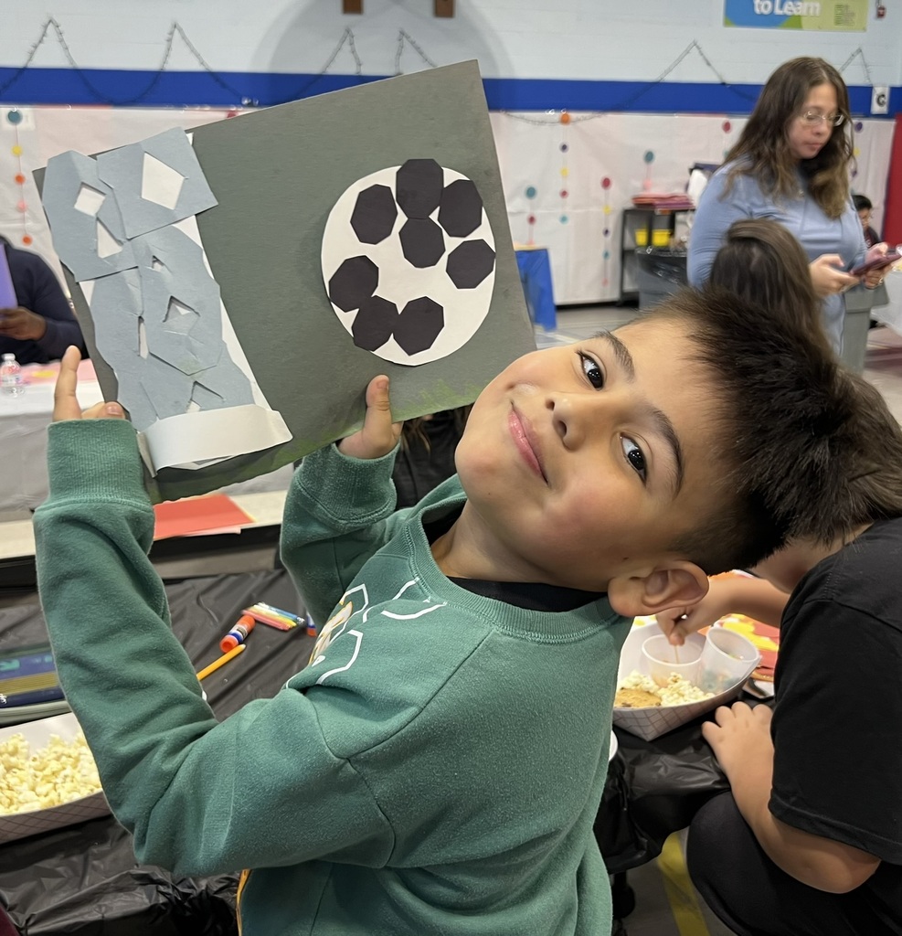 A smiling child in a green sweatshirt proudly holds up a gray paper collage of a soccer ball and geometric shapes in a busy classroom.
