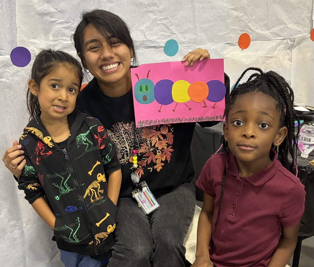 A smiling woman poses with two young girls, holding a pink artwork of colorful caterpillars; paper circles decorate the wall behind them.