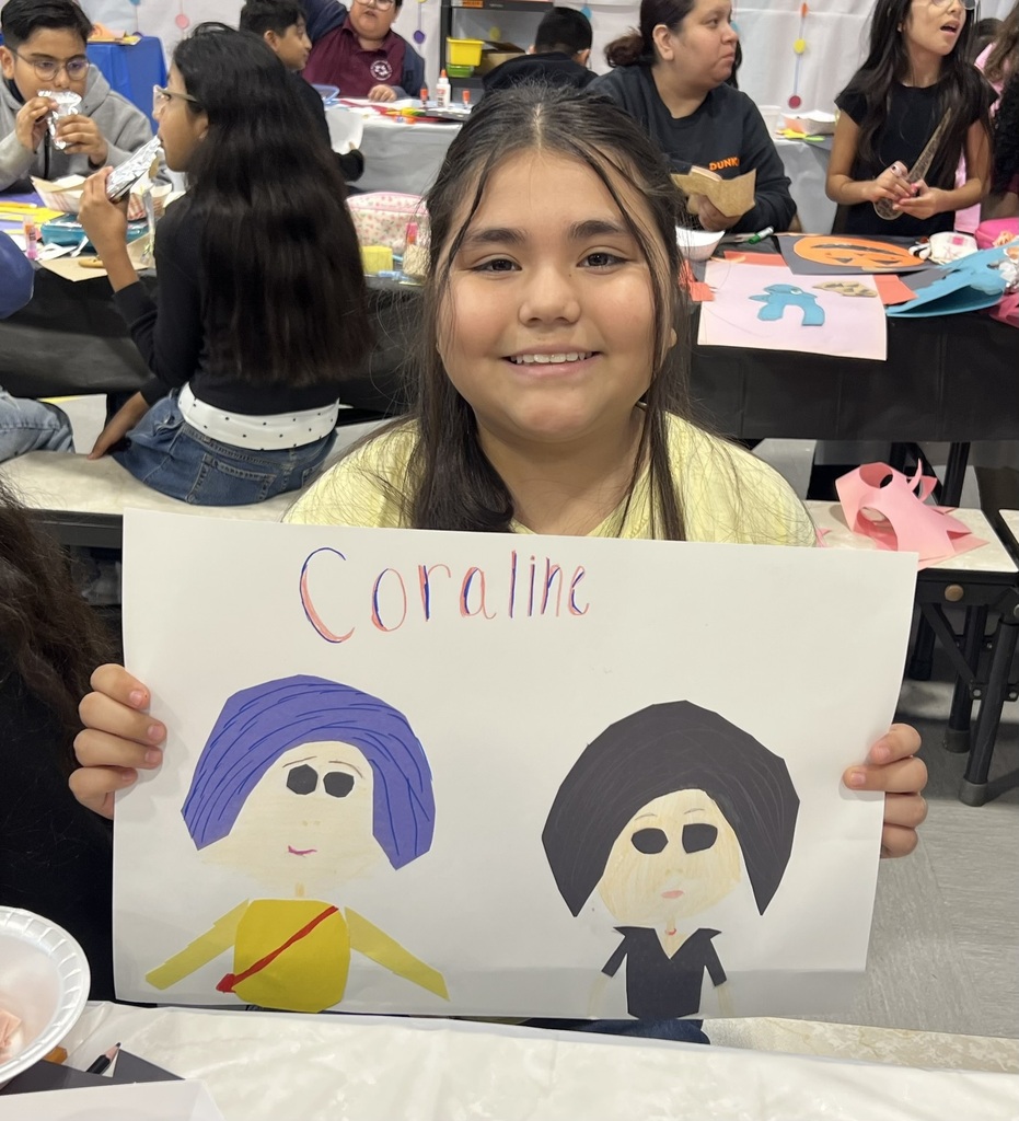 A smiling student holds a handmade Coraline poster with two colorful character drawings, while classmates work on crafts behind her in a lively classroom.