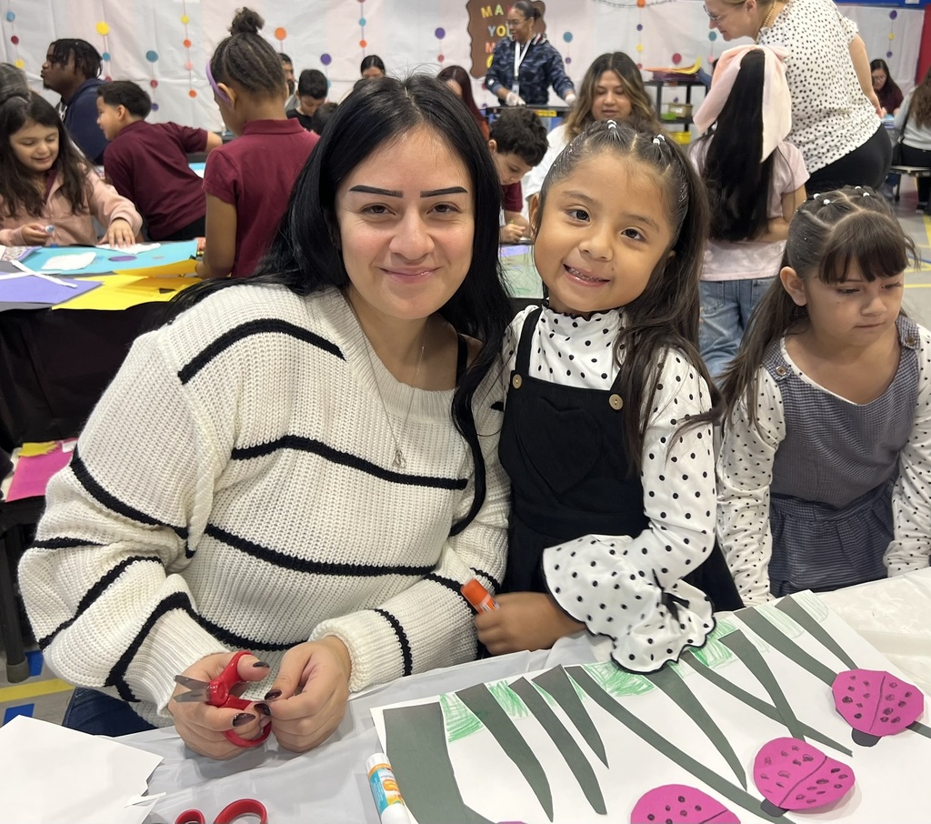 A smiling woman and young girl pose beside a colorful classroom art project as children craft at tables in a lively, welcoming school setting.