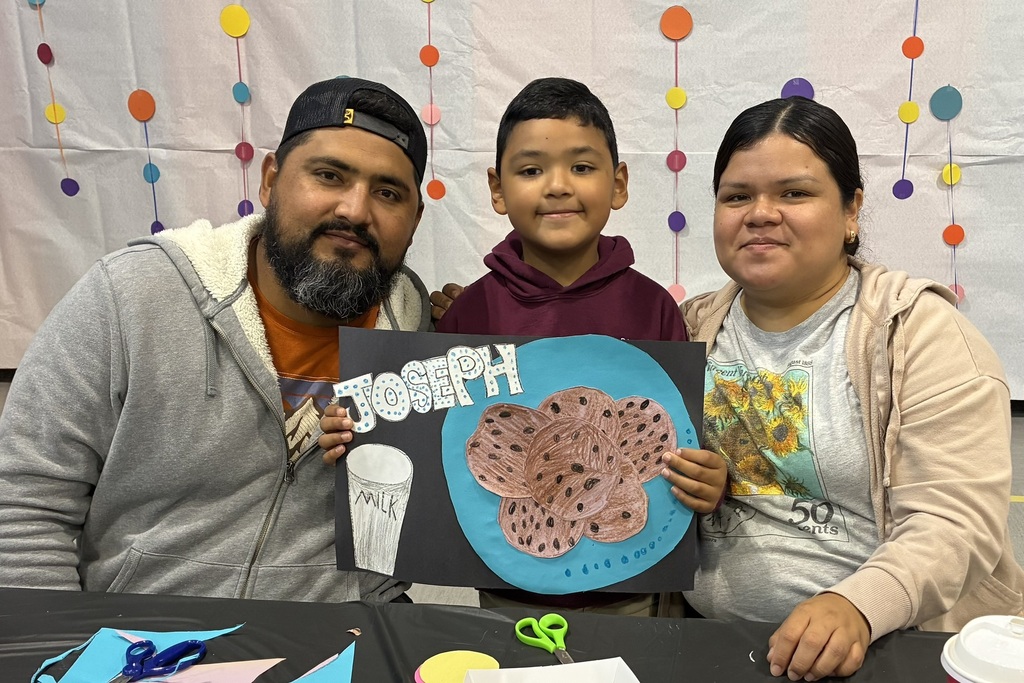 A smiling family poses in a decorated classroom as a boy holds a blue poster showing chocolate-chip cookies and a glass of milk.