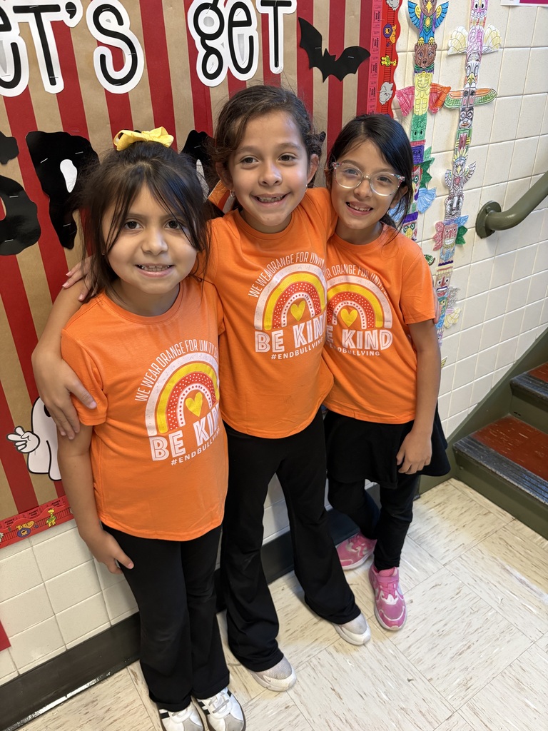 Three smiling schoolchildren pose arm-in-arm in matching orange “Be Kind” shirts, black pants, and colorful shoes beside a red-and-white classroom kindness display.