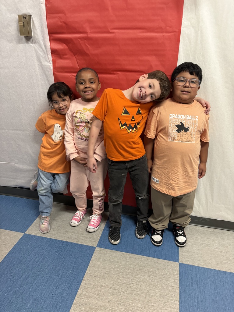 Four children pose together in Halloween-themed orange shirts before a red-and-white backdrop, smiling and leaning close on a blue-and-gray tiled floor.