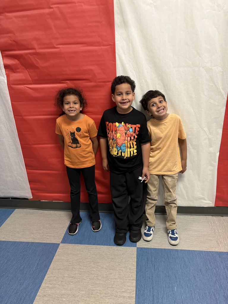 Three smiling children pose together against a red-and-white backdrop on a blue and gray tiled floor, creating a cheerful, playful school-event photo.