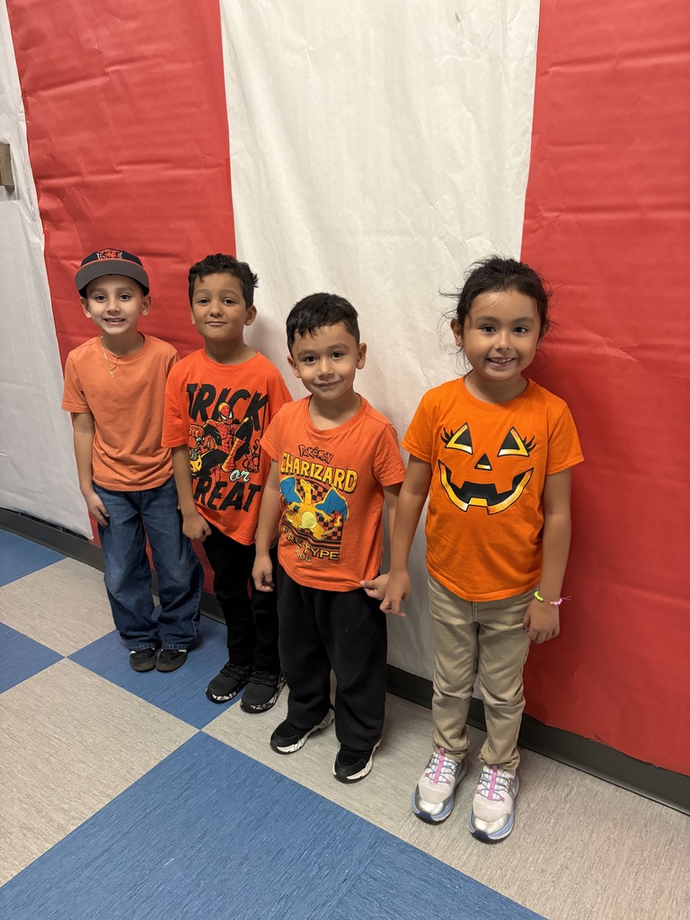 Four smiling children in orange Halloween shirts and costumes stand together against a red-and-white backdrop in a school hallway, creating a cheerful mood.