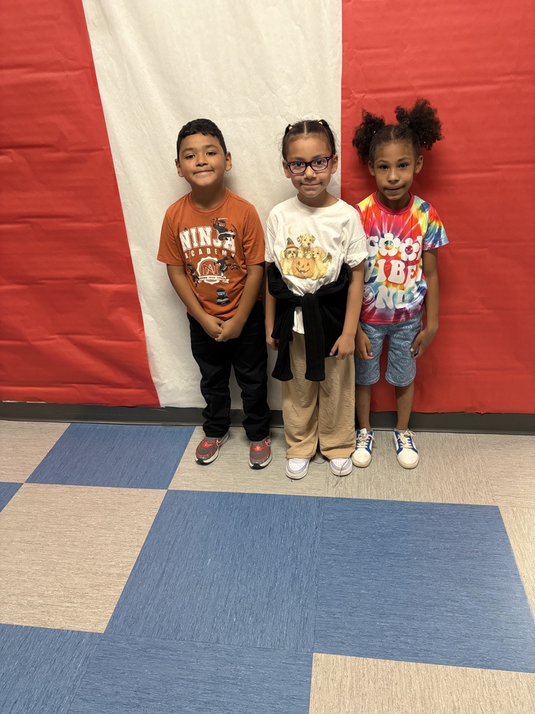 Three young children pose together in a school gym, smiling in colorful casual clothes before a large red-and-white wall, with blue and tan floor tiles.