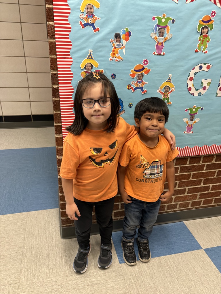 Two children in orange Halloween shirts stand arm in arm before a turquoise bulletin board decorated with colorful clowns and a large letter G, smiling warmly.