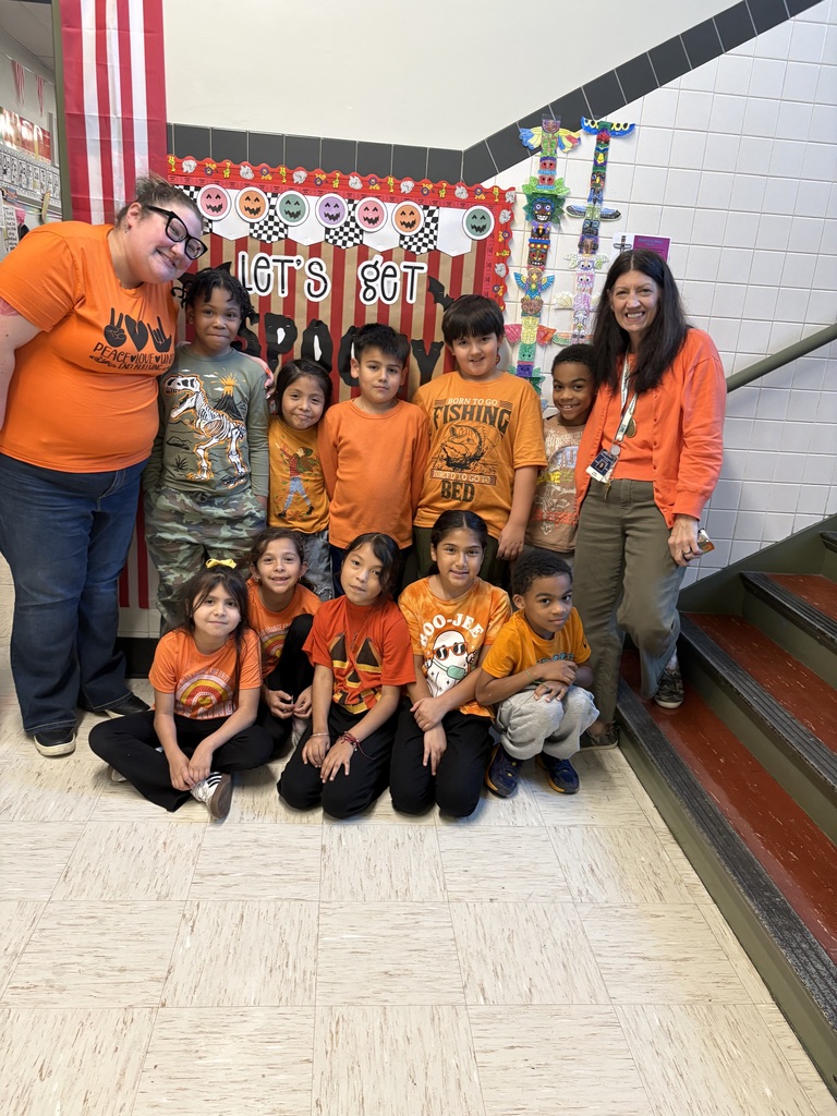 Teachers and children in orange shirts smile for a group photo before a festive Halloween display with pumpkins and colorful decorations in a school hallway.