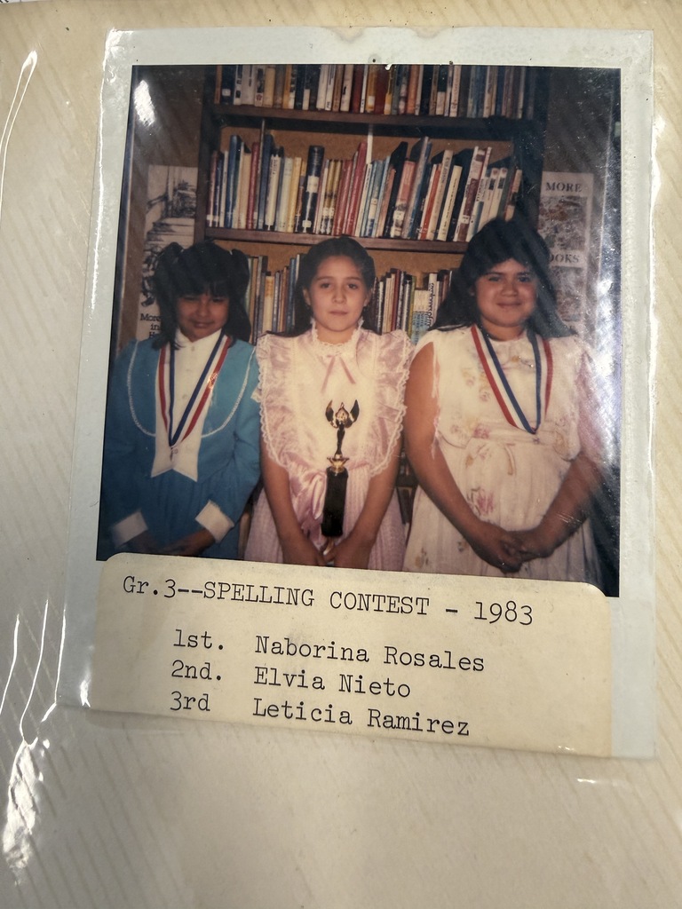 A worn 1983 spelling contest photo shows three girls posing with a trophy before bookshelves; a caption lists their places, capturing a proud, nostalgic moment.