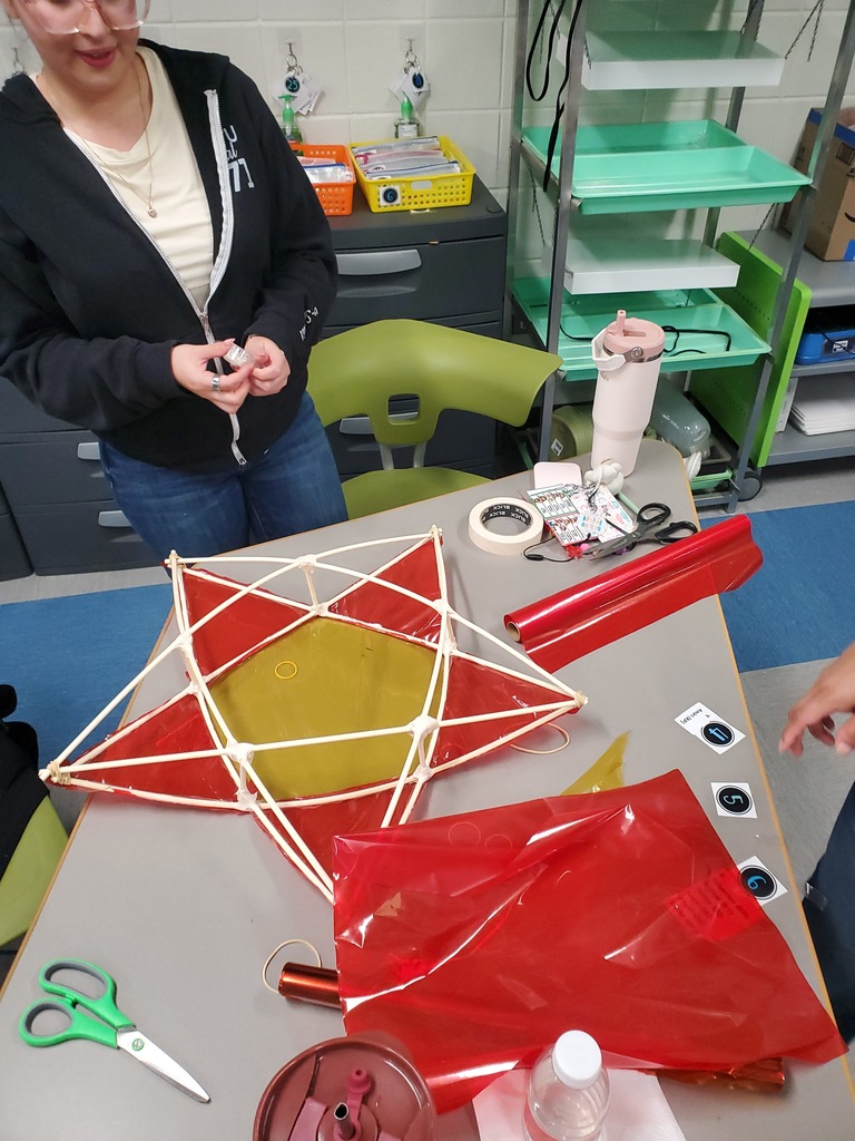 A workshop participant demonstrates a red-and-white geometric kite on a table with red plastic sheets, scissors, and craft supplies in a bright lab.