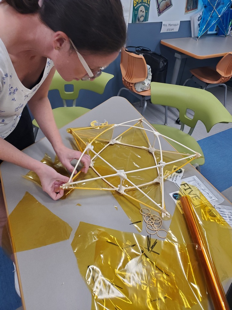 A student carefully assembles a white geometric frame over shiny gold foil on a classroom table, focused amid colorful chairs and art supplies.
