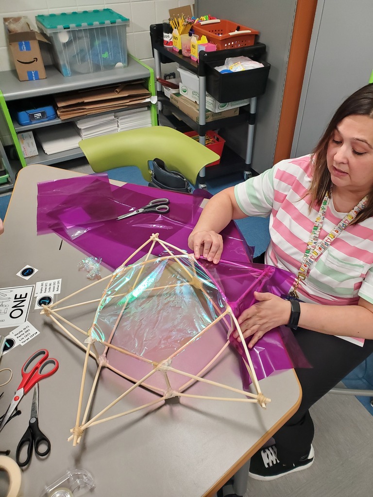 A woman in a craft room assembles a bamboo-frame sculpture covered in iridescent purple and clear film, with scissors and supplies on the table.