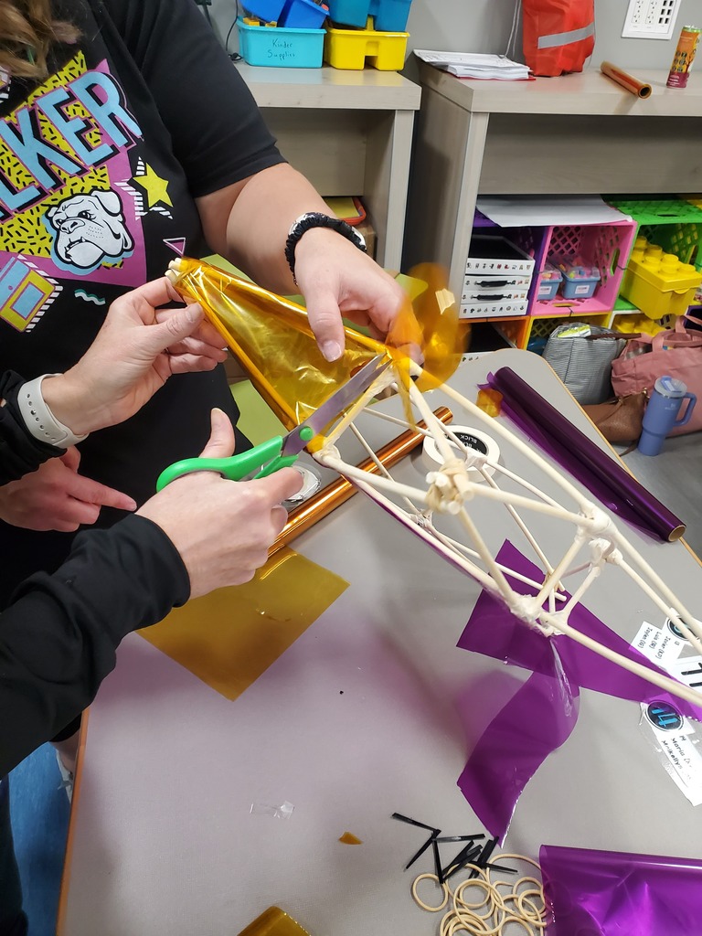 Hands craft a kite in a colorful classroom, trimming shiny gold foil with green scissors around a wooden frame; purple tails and rubber bands lie on the table.