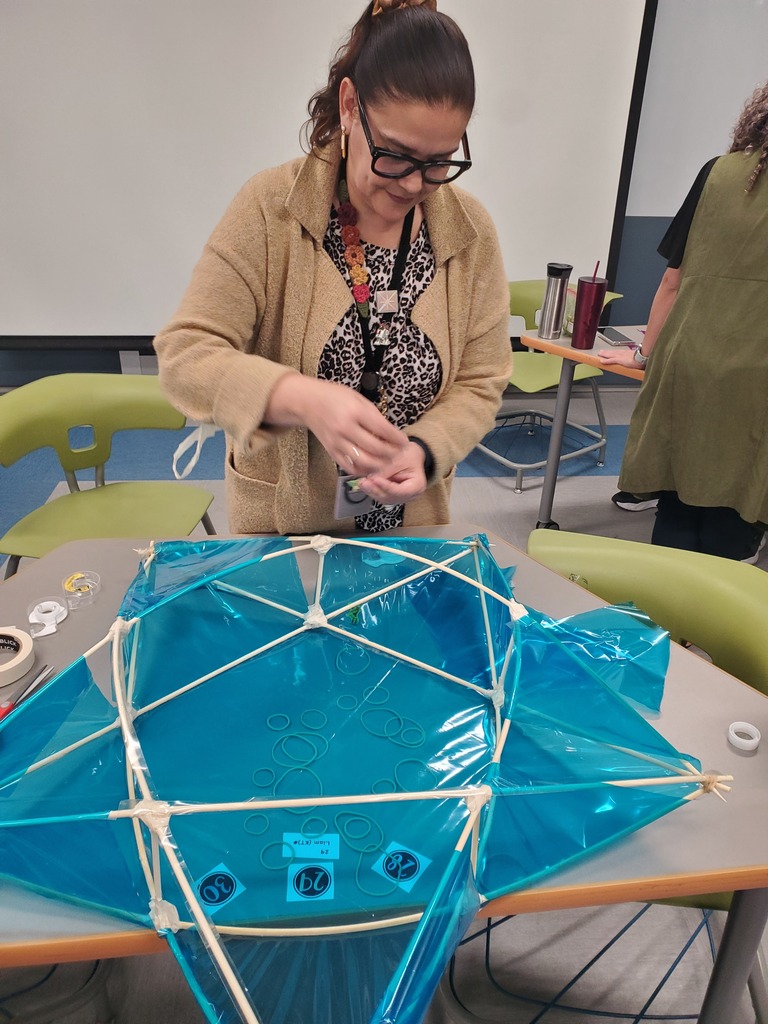 A woman in a classroom carefully assembles a geometric structure of blue plastic and wooden sticks on a table, creating a focused, collaborative mood.