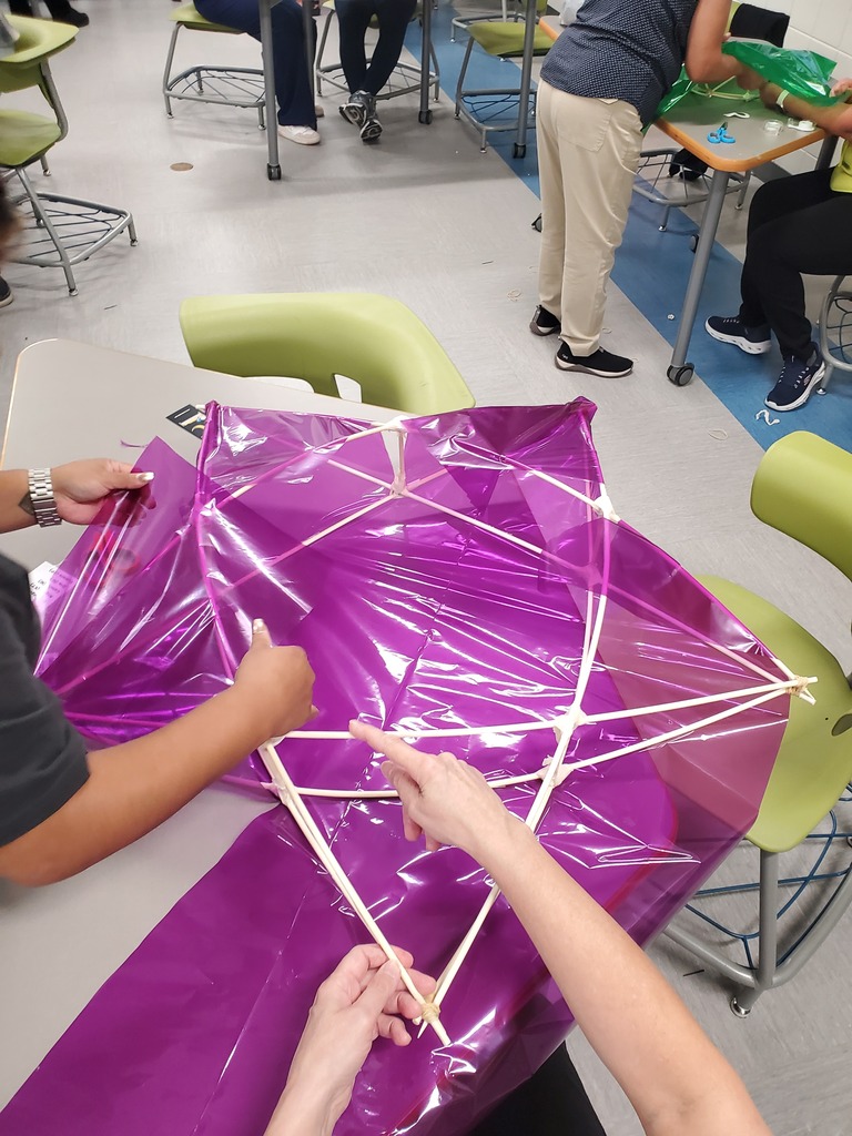 Several people assemble a large purple plastic kite, fastening thin wooden spars in a classroom; the scene conveys focused teamwork.
