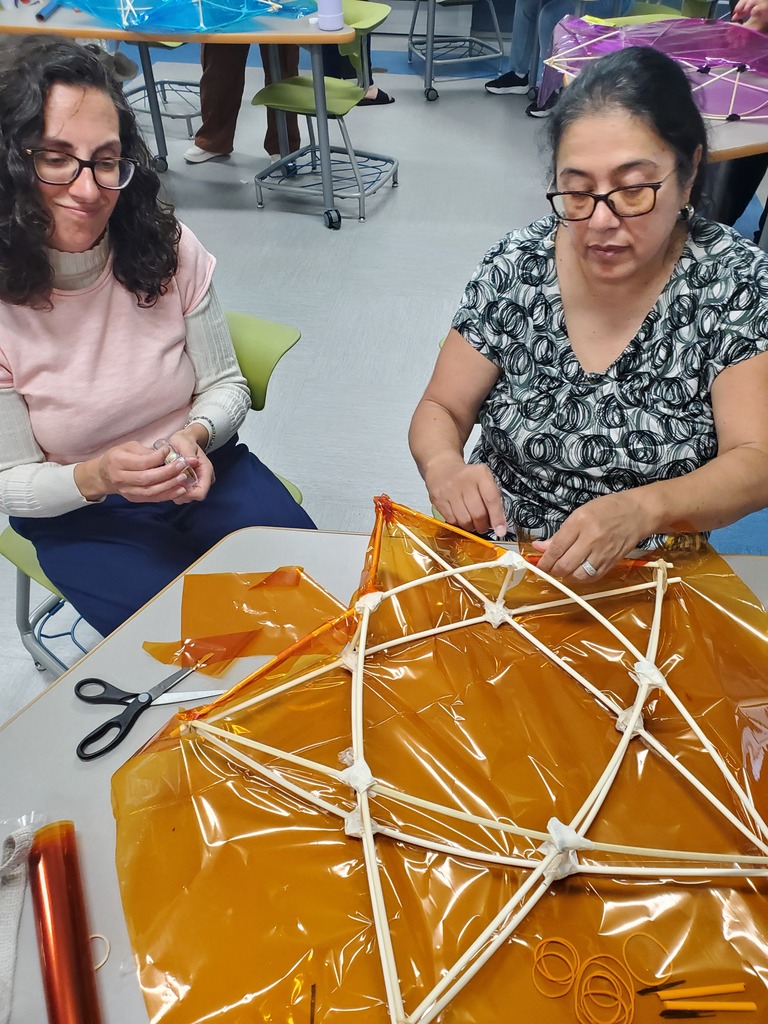Two women work together at a table, assembling a geometric structure from white rods and shiny orange plastic. Their focused expressions create a calm, collaborative workshop atmosphere.
