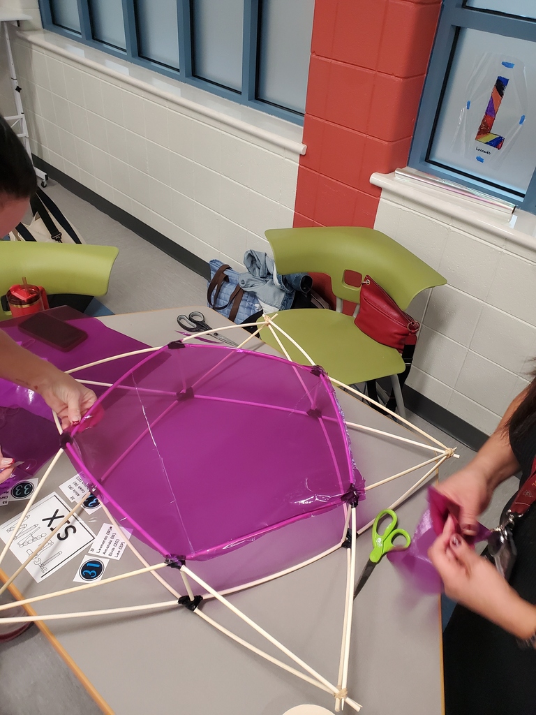 Students assemble a large purple kite-like frame with wooden rods and string on a classroom table, sharing a focused, hands-on craft activity.