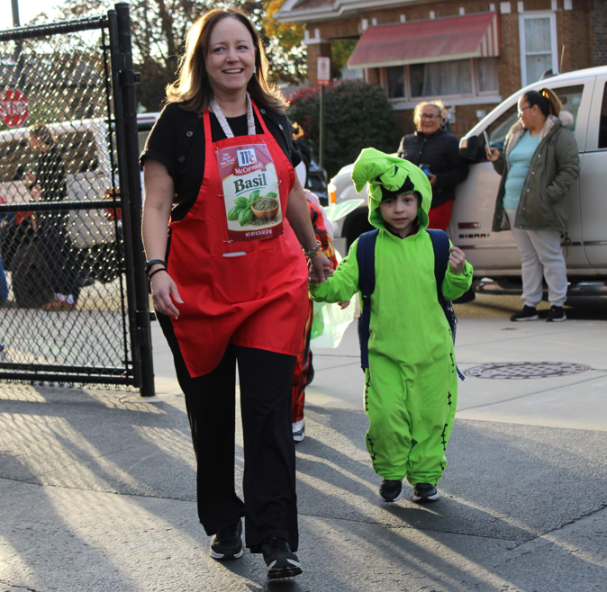A smiling woman in a red apron walks hand in hand with a child in a bright green dinosaur costume at a neighborhood event.