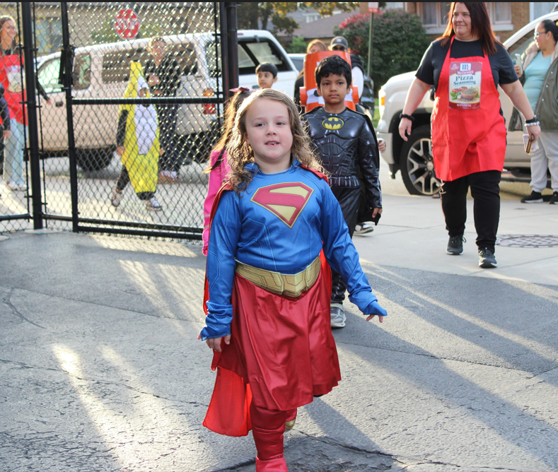 A smiling child in a Supergirl costume walks toward the camera at a cheerful outdoor Halloween event, with other costumed children and adults behind her.