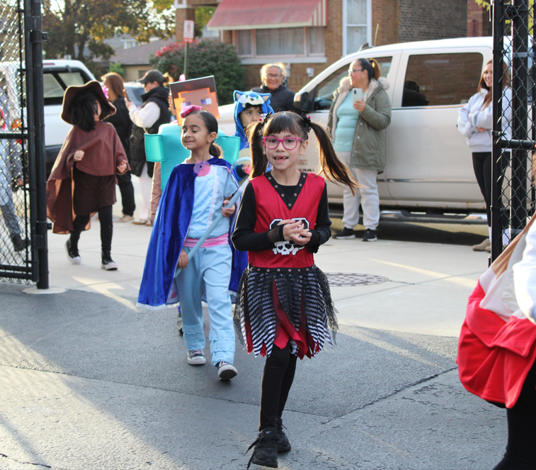 Children in colorful Halloween costumes parade through a school gate, led by a smiling girl in pink glasses and a black-and-red outfit, as adults watch nearby.