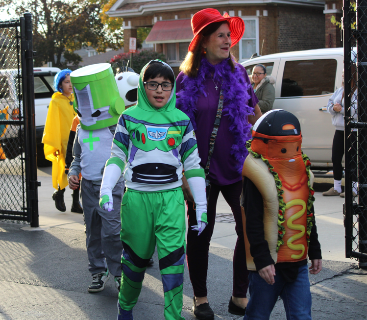 A group in Halloween costumes walks through a schoolyard gate: a child dressed as Buzz Lightyear leads, with a costumed adult and other children behind. The scene feels cheerful and festive.
