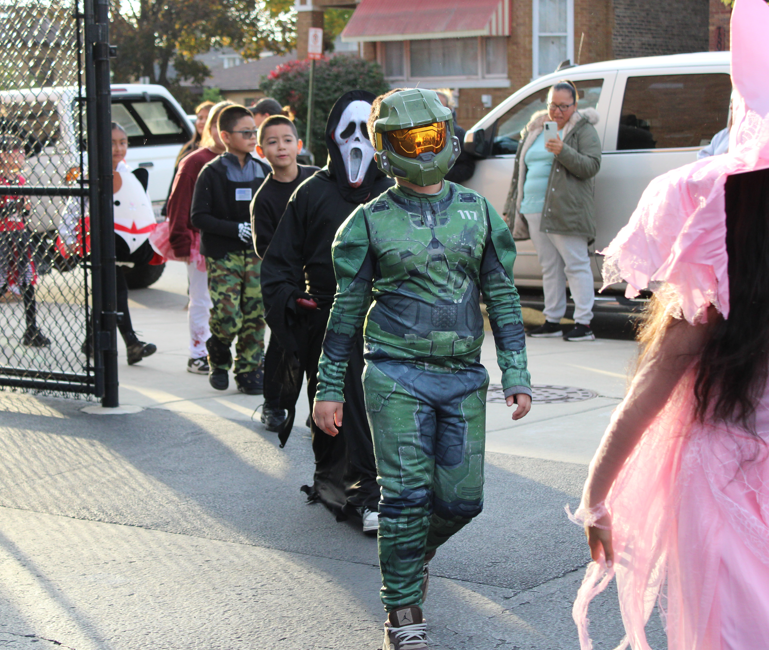 People in Halloween costumes, including a green-armored sci-fi soldier and Ghostface, walk along a sunny street as onlookers watch.