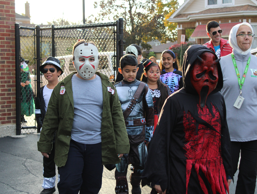 Children in Halloween costumes, including Jason Voorhees and a red-masked figure, walk past a chain-link gate with adults nearby; festive, playful mood.