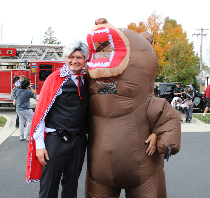 A smiling person in a red cape and white wig poses beside a bulky brown monster costume, with a fire truck at an outdoor Halloween event.
