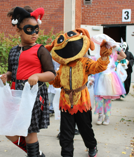 Children in Halloween costumes collect candy outside a brick school, including a masked girl and a grinning orange monster mascot; festive, playful mood.