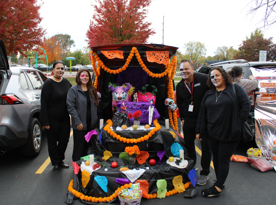 Four adults pose beside a colorful Halloween Día de los Muertos altar display with orange marigold garlands, skulls, candles, and flowers in a parking lot.