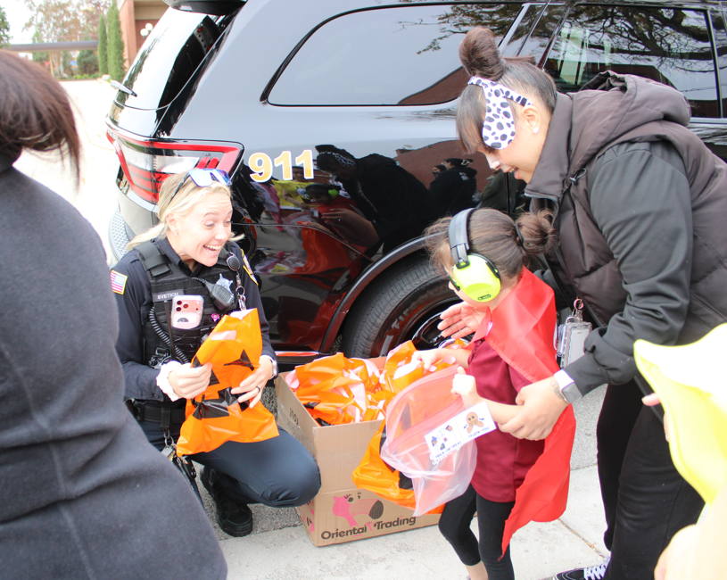 A uniformed police officer kneels beside children in Halloween costumes, handing out orange candy bags near a black patrol SUV in a cheerful community event.