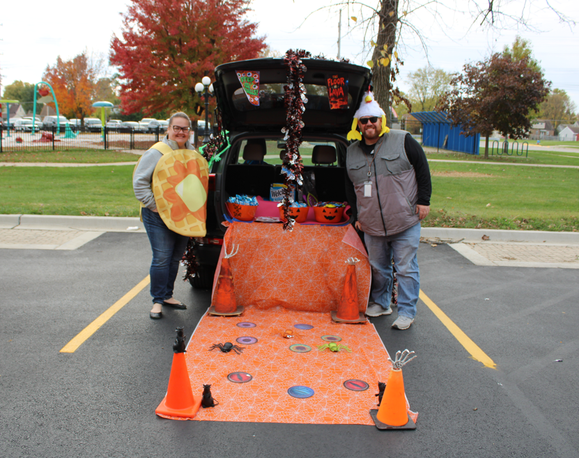 Two adults pose beside a Halloween-decorated car trunk with an orange game ramp and traffic cones in a fall parking lot, creating a playful mood.
