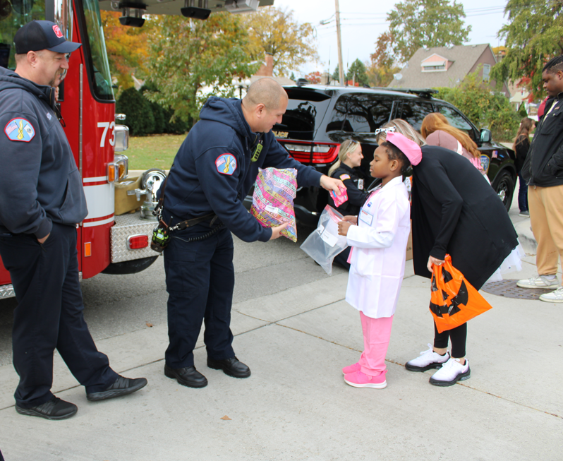 A firefighter hands a colorful Halloween treat bag to a young girl in a pink costume beside a fire truck, as neighbors watch warmly.