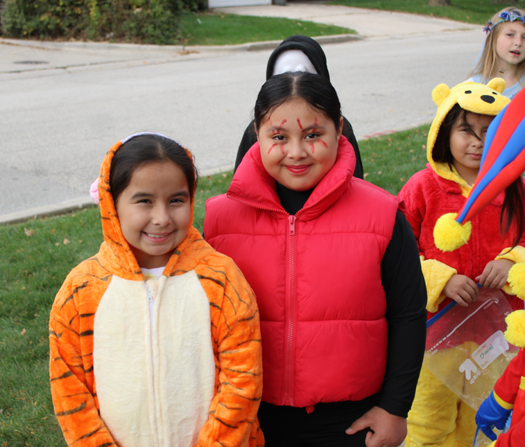 Children in colorful Halloween costumes, including tiger and bear outfits, smile at a community gathering; a child in a red vest wears face paint.