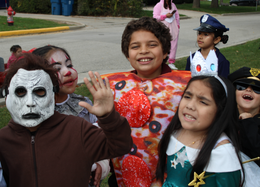 Children in Halloween costumes gather outdoors; a smiling boy in a pizza costume waves beside a masked figure, police officer, and princess.