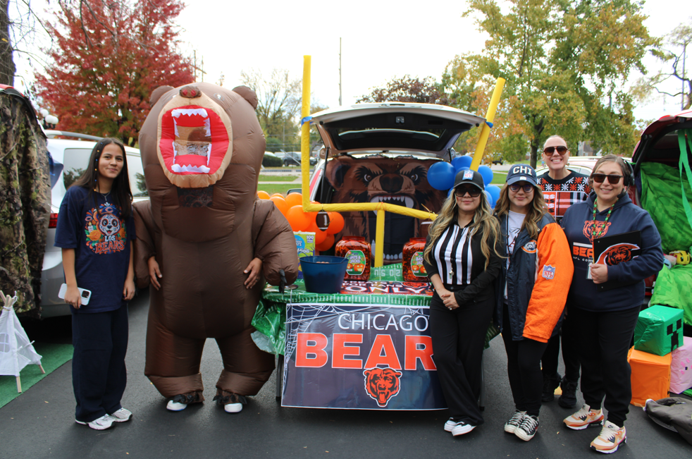 Chicago Bears fans pose at a fall tailgate beside a roaring bear mascot and decorated car, smiling among team flags and gear in a festive scene.
