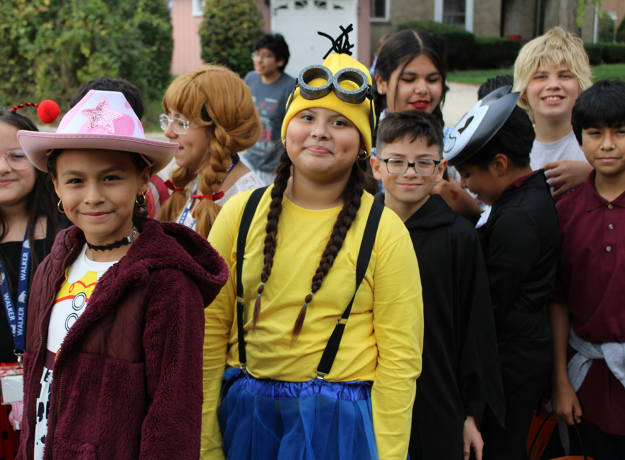 Children in Halloween costumes gather outdoors, including a girl dressed as a Minion in yellow and blue and another in a pink cowboy hat; the group looks cheerful and playful.