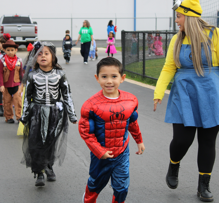 Children in Spider-Man, skeleton, and cowboy costumes walk outdoors at a Halloween event as a smiling adult in a yellow hat and blue dress guides them.
