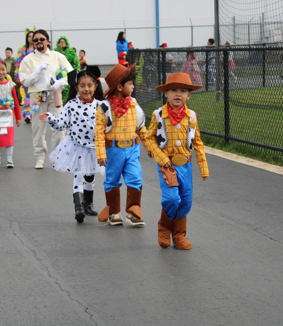 Children parade outdoors in colorful Halloween costumes, led by two children dressed as cowboys and a girl in a spotted cow outfit; the mood is cheerful.