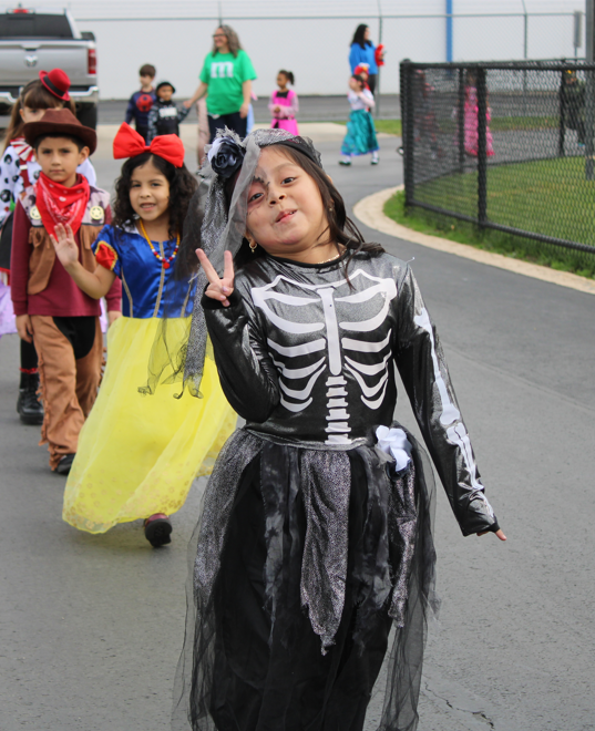 A cheerful child in a black skeleton costume poses with a peace sign at a school Halloween parade, with costumed classmates following behind.
