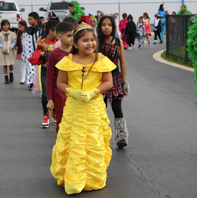 A smiling child in a bright yellow princess gown leads a line of children in Halloween costumes along a school roadway, creating a cheerful parade scene.