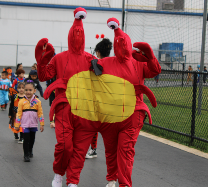 Two adults in a bright red crab costume pose with raised claws as children in colorful outfits walk behind them; a cheerful school parade outdoors.