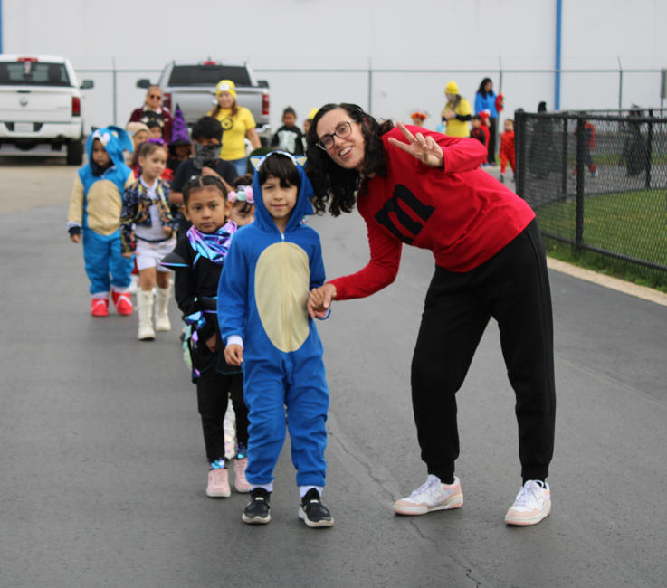 A smiling woman in a red top poses with a child in a blue dinosaur costume as a line of children follows outdoors, creating a cheerful, playful mood.
