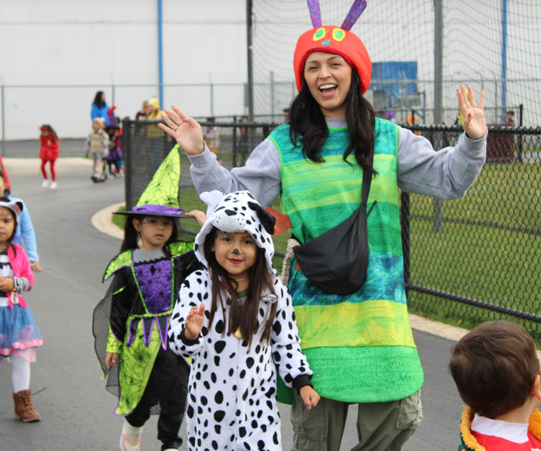 A smiling woman in a bright green costume and red hat poses with children in Halloween costumes at an outdoor event, creating a cheerful, playful mood.