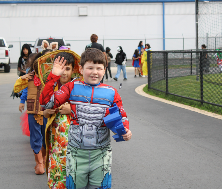 A smiling child in a blue-and-red superhero costume poses with a toy blaster while a child in a colorful costume waves behind him at an outdoor event.