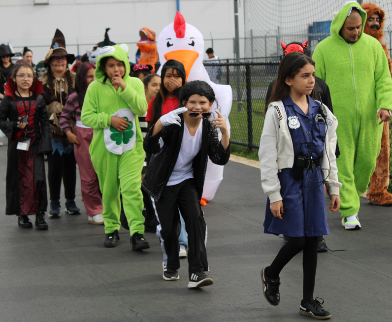 Children in Halloween costumes walk outdoors, led by a girl dressed as a doctor, with a chicken mascot and colorful animal and witch costumes behind her.