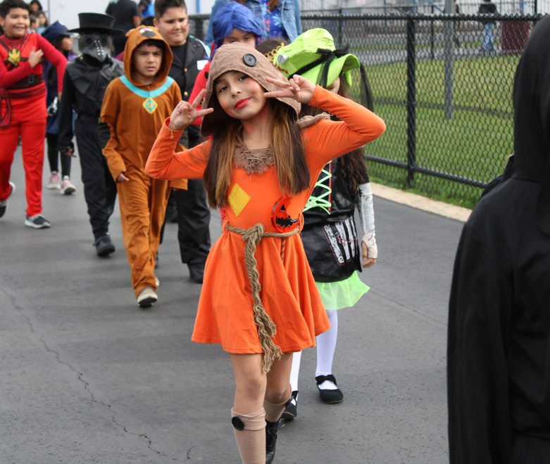 A smiling girl in an orange Halloween costume poses with peace signs during a school parade, surrounded by children in colorful costumes. The mood is playful and festive.
